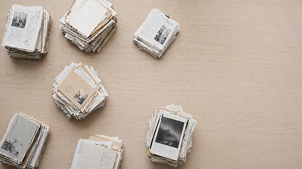 Mehrere voneinander getrennte Stapel aus Papierdokumenten auf einem großen hellen Holztisch von oben fotografiert.