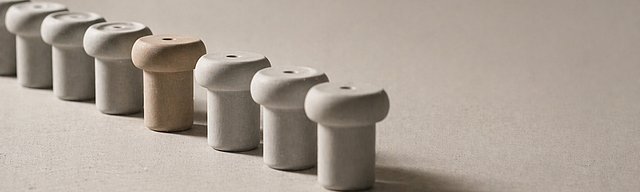Reihe identischer heller Keramik-Objekte im Studiolicht, bei der ein einzelnes Stück leicht nach vorne versetzt und wärmer getönt ist.