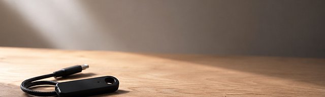 Mattschwarzes Hardware-Security-Token mit gewickeltem USB-C-Kabel auf einer Eichenholz-Oberfläche, weiches Tageslicht von links — Metapher für die Permissions- und Identitäts-Boundary, die Project Arc und OpenShell für autonome Agenten schaffen.