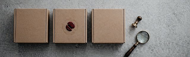 Drei identische Kraftpapier-Versandkartons in einer Reihe auf Beton, der mittlere mit gebrochenem dunkelrotem Wachssiegel, daneben ein Messingstempel und eine Lupe.