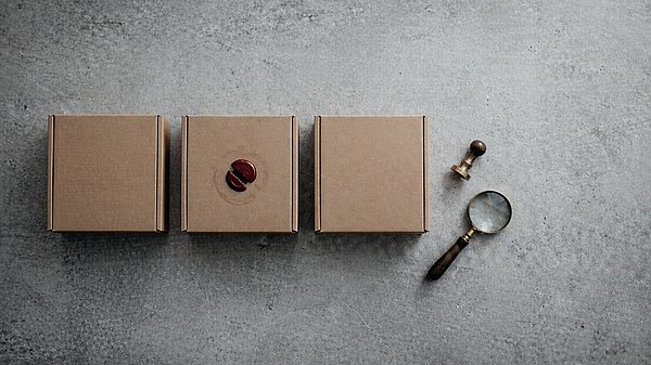 Drei identische Kraftpapier-Versandkartons in einer Reihe auf Beton, der mittlere mit gebrochenem dunkelrotem Wachssiegel, daneben ein Messingstempel und eine Lupe.