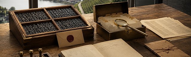 Walnuss-Schreibtisch mit antiquem Setzkasten (vier Fächer voller Bleilettern), geöffneter Brass-Karteikassette mit Kraftpaper-Tags, messingfarbener Lupe und einem aufrecht gestellten Oxblood-Wachssiegel-Etikett; im Hintergrund die Glasfront eines modernen Moselhauses mit sonnigem Weinberg-Hang — visuelle Metapher für die vier Schichten eines AI-ready CMS: strukturierte Inhalte, semantische Auslieferung, Agent-Interaktion und Vertrauen.