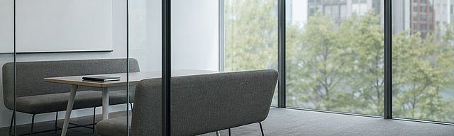 Aufgeräumter moderner Besprechungsraum hinter einer Glaswand mit leerer Whiteboard-Wand und zwei gegenüberstehenden Sitzbänken.
