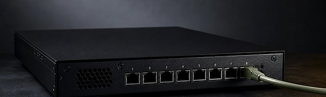 Mattschwarze Server-Edge-Box mit acht Netzwerkports — nur einer verbunden mit einem sage-grünen Patch-Kabel, sieben absichtlich offen, kühles Studiolicht von links — Metapher für deterministische Isolation als Security-Default.