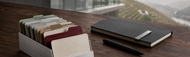 Brushed-Aluminium-Karten-Halter mit gefächerten Pastell-Karten auf Walnuss-Schreibtisch vor heller Mosel-Glasfront; eine Karte in Oxblood ist nach vorn gezogen, daneben ein geschlossenes mattschwarzes Notizbuch.