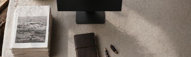 Aufgeräumter Redaktions-Arbeitsplatz von oben mit gestapelten gedruckten Magazinen neben einem ausgeschalteten Monitor.