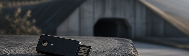 Hardware-Security-Token auf verwittertem Beton-Sockel, im Hintergrund weich gezeichneter Cyber-Bunker-Eingang in der Mosel-Steillage, kühles Morgenlicht.