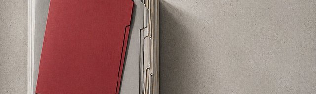 Präzise gestapelte graue Aktenmappen von oben fotografiert, eine rote Mappe hebt sich deutlich ab.