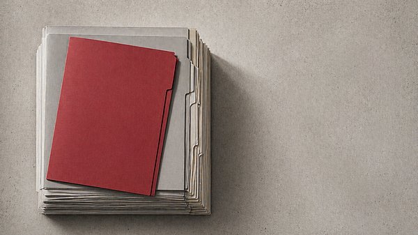 Präzise gestapelte graue Aktenmappen von oben fotografiert, eine rote Mappe hebt sich deutlich ab."