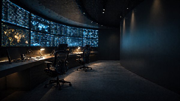 Abgedunkelter Leitstand bei Nacht mit zwei leeren Bürostühlen vor einer Wand aus leuchtenden Monitoren.